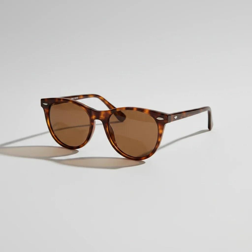 A pair of classic-style sunglasses with brown lenses and dark tortoiseshell frames, resting on a white table next to a sandwich on a plate and an open book.