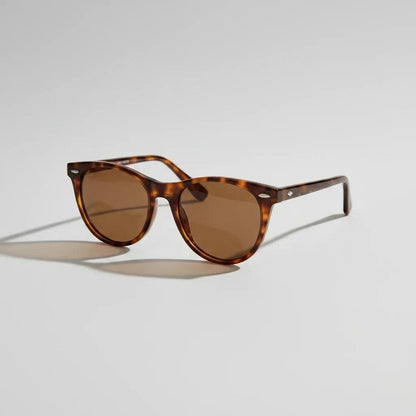 A pair of classic-style sunglasses with brown lenses and dark tortoiseshell frames, resting on a white table next to a sandwich on a plate and an open book.